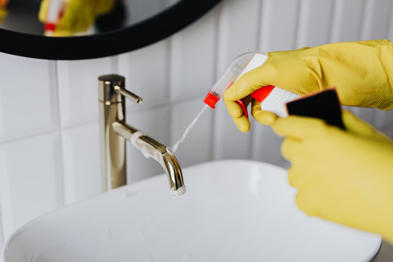 Anonymous housekeeper above, wearing yellow rubber gloves, sprays cleaner onto the chrome bathroom faucet.